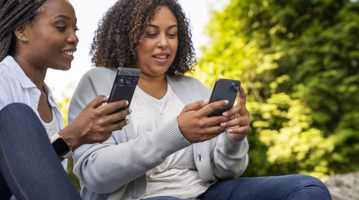 Two women sit outside together playing on their smartphones. 